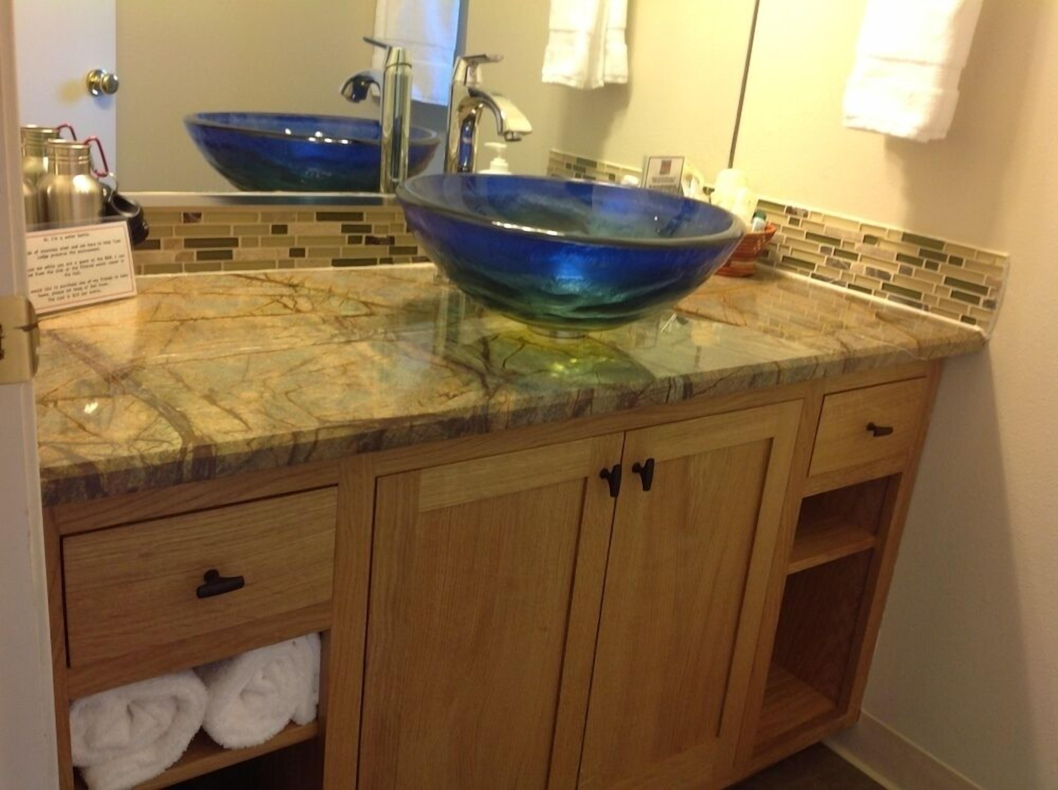A bathroom with a large blue glass bowl sink, wooden cabinets, and a tiled backsplash.