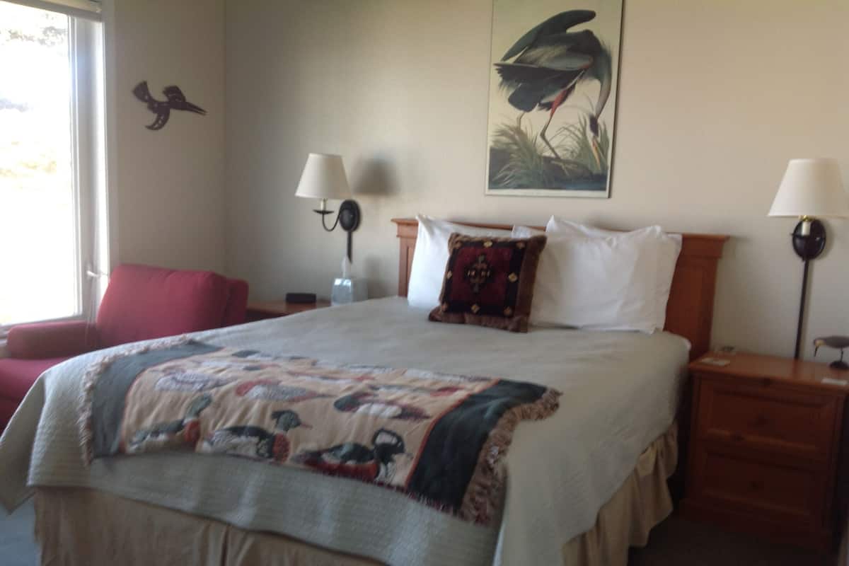 A neatly made bed with a quilt, two bedside lamps, a red chair, a wall-mounted bird sculpture, and a painting of a bird.