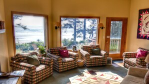 A cozy living room with plaid furniture, large windows offering a view of the ocean, and a door leading to a balcony. - Tyee Lodge Oceanfront B&B (Newport)