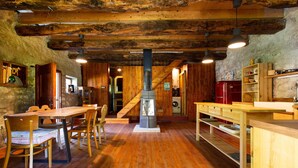 Ein rustikales Blockhaus-Interieur mit einem Holzesstzimmer, Stühlen, einem zentralen Holzofen und einer Küchenzeile mit Holzregalen und einem roten Kühlschrank.