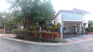 Un edifici residencial amb entrada tancada per una reixa, un camí d'accés pavimentat i una zona de jardí ben cuidada amb diferents plantes.