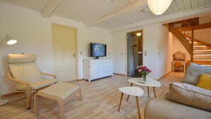 Una sala de estar acogedora con piso de madera, un sofá blanco, un sillón, una mesita con un florero, un televisor sobre un mueble blanco y una escalera que conduce a un nivel superior.
