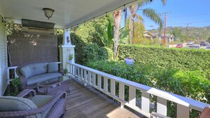 A covered porch with wicker furniture, a white railing, and a view of a lush green landscape with a road and houses in the background. - Downtown Craftsman Bungalow (Santa Barbara)