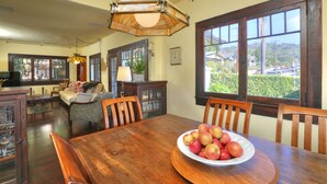 A dining area with a wooden table, chairs, and a bowl of apples. There is a window with a view of a parking lot and greenery. - Downtown Craftsman Bungalow (Santa Barbara)