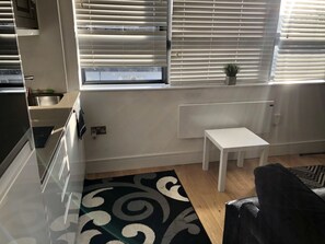 A compact kitchen with a built-in oven, a small sink, and a window with blinds. There is a white table and a black chair in the room.