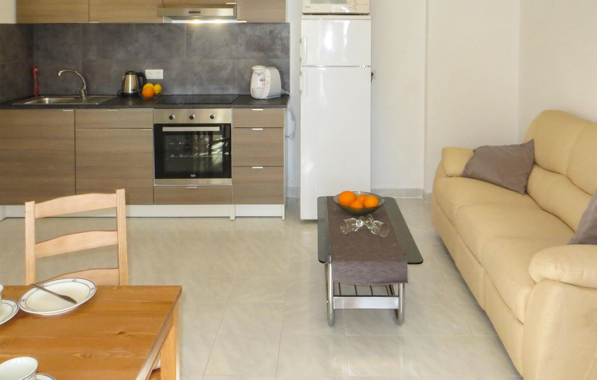 A compact kitchen with a built-in oven, a refrigerator, and a microwave. There's a dining table with plates and a bowl of fruit. A sofa is in the living area.