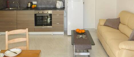 A compact kitchen with a built-in oven, a refrigerator, and a microwave. There's a dining table with plates and a bowl of fruit. A sofa is in the living area.