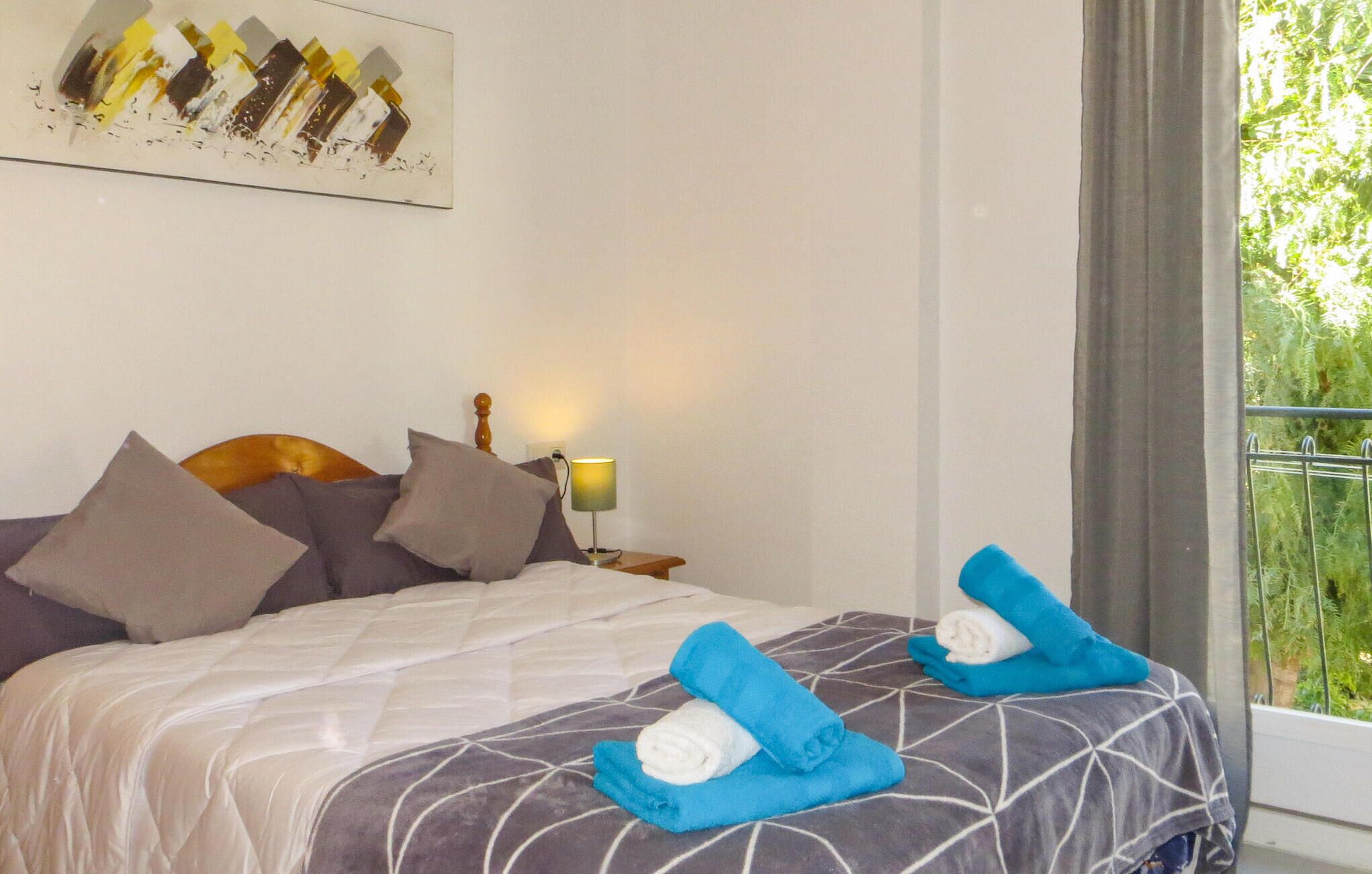A neatly made bed with a patterned bedspread, two pillows, and two rolled towels on the bed.