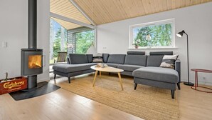 Una sala de estar moderna con estufa de leña empotrada, sofá seccional gris y un ventanal con vistas a árboles.