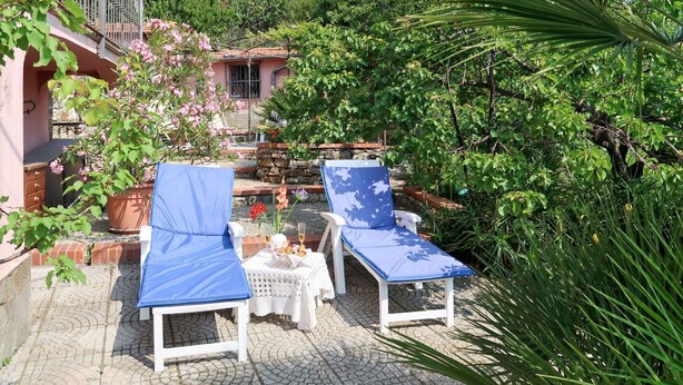Uma área de pátio com duas poltronas brancas cobertas por almofadas azuis, uma mesa pequena com uma cesta de lanches e um muro de pedra ao fundo.