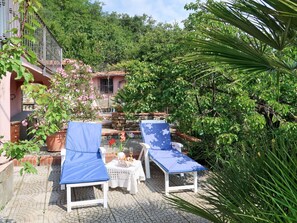 Un patio con dos sillones blancos cubiertos de cojines azules, una mesita con una cesta de aperitivos y un muro de piedra al fondo.