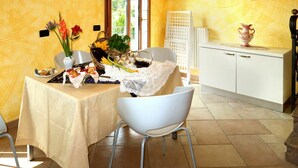 Uma área de refeições com uma mesa posta para dois, uma cadeira branca, um armário branco e um vaso com flores.
