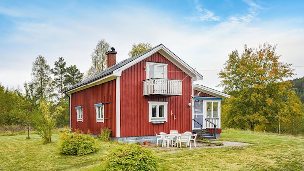 Una casa de madera roja con balcón, ventanas blancas y un pequeño porche con muebles de exterior.