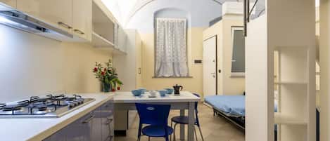 A compact kitchen with a gas stove, a dining table with chairs, a small window with curtains, and a bed in the adjacent room.