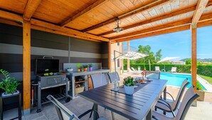 Une terrasse extérieure équipée d’un barbecue, d’une table à manger, de chaises et offrant une vue sur une piscine et un espace verdoyant.