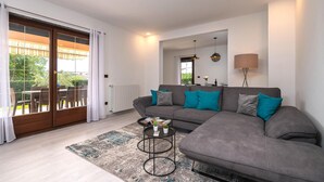 Un salon moderne avec un canapé gris, des coussins turquoise, une petite table basse ronde et une grande fenêtre donnant sur l’extérieur.