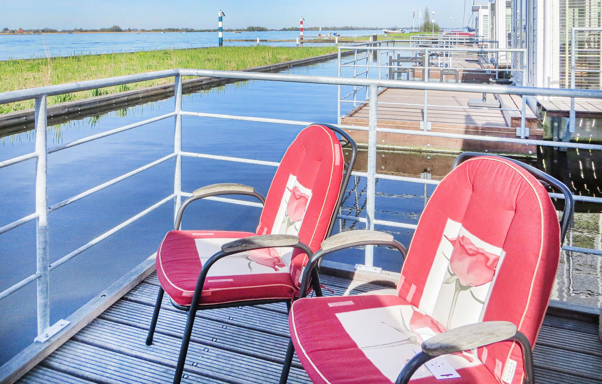 Two red chairs with floral cushions on a metal deck overlooking a canal with a waterway and a distant shoreline.