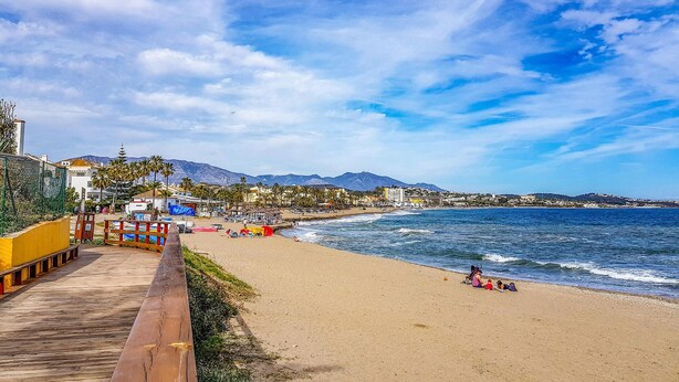 Una playa con un paseo de madera, palmeras y edificios al fondo.
