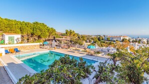 Zona de piscina con sillones de descanso y sombrillas, rodeada de vegetación y edificios al fondo.