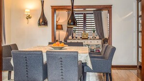Uma sala de jantar com uma mesa posta para seis pessoas, rodeada por seis cadeiras cinza estofadas com frente abotoada. Há duas luminárias fixadas na parede acima da mesa, e um aparador de madeira com objetos decorativos e um relógio ao fundo.
