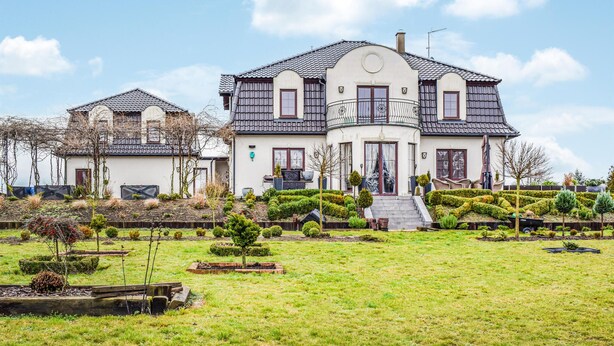 Uma casa grande de dois andares com um jardim bem cuidado, uma pequena escadaria para a entrada e um céu limpo.
