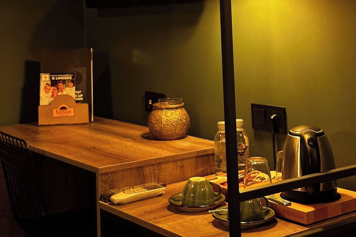 Una habitación con poca luz, una mesa de madera, un televisor montado en la pared, una lámpara y varios objetos sobre la mesa, incluyendo un control remoto, una tetera y una botella.