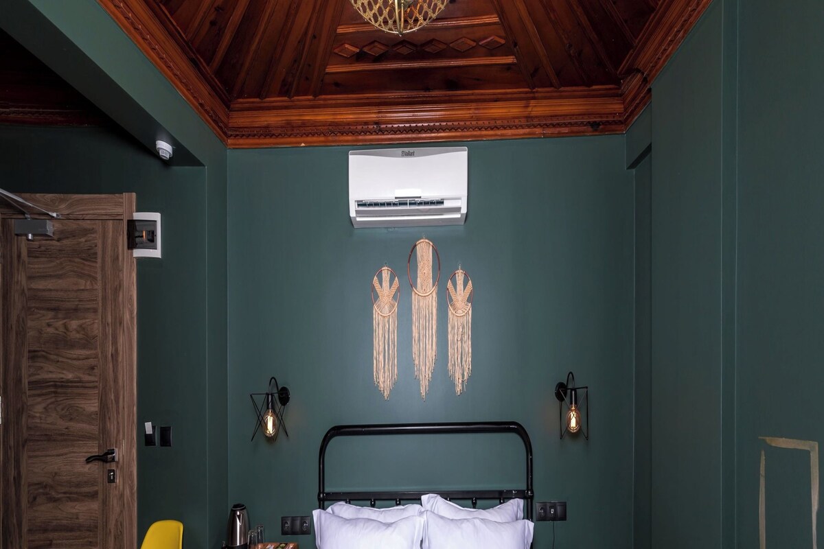 A room with a bed, a yellow chair, a wooden door, a wall-mounted air conditioner, and a ceiling light fixture.