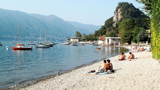 Une plage de galets où des gens se prélassent au soleil, des bateaux sur l’eau et des montagnes en arrière-plan.
