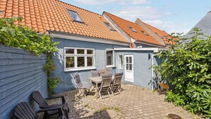 Een blauw huis met een pannendak, een kleine tuin met een tafel en stoelen, en een houten hek.
