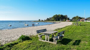 Ein Strand mit einer Holzbank und einem Picknicktisch, Booten auf dem Wasser und Häusern im Hintergrund.