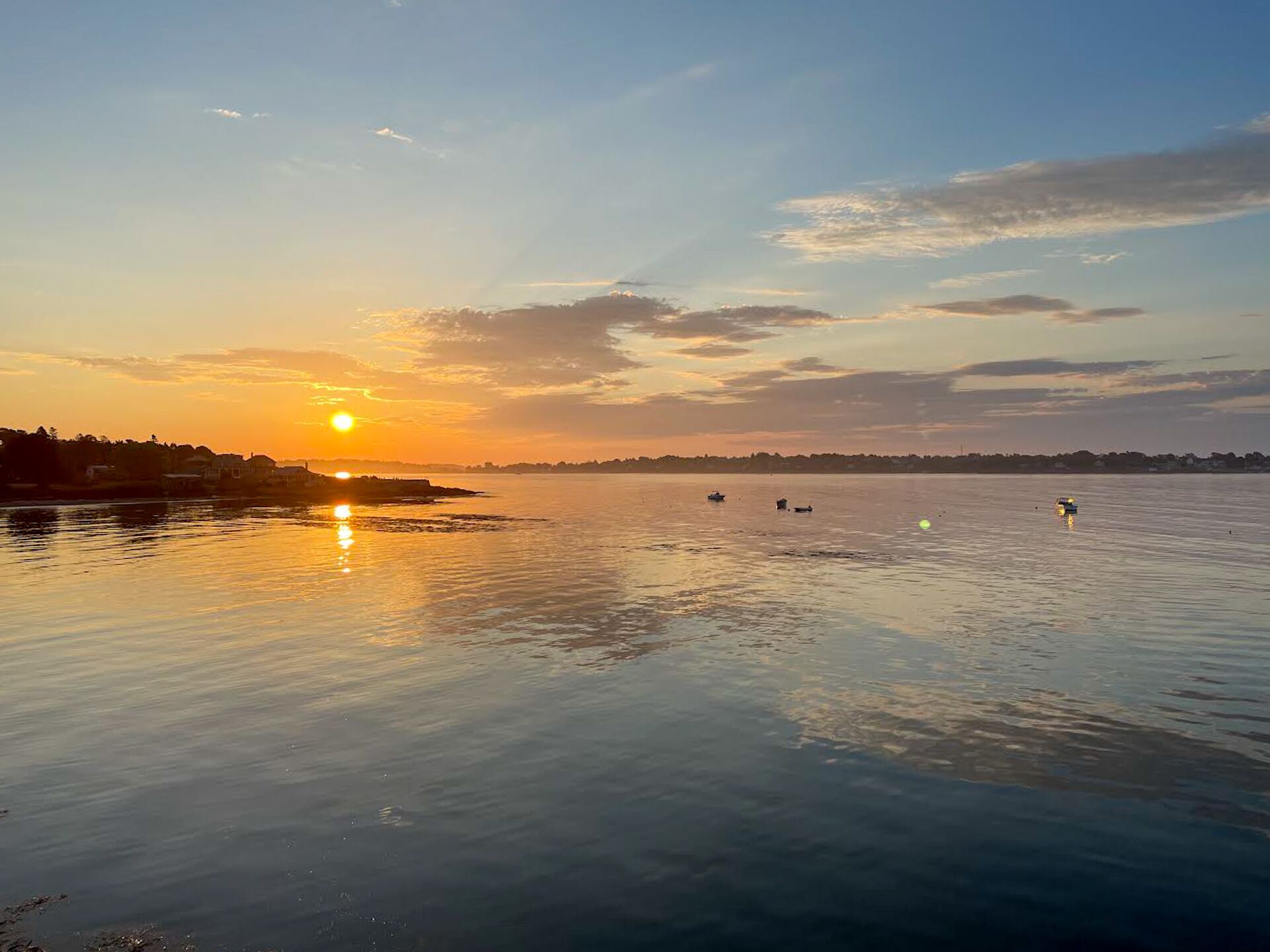 Une étendue d’eau calme, le soleil se couchant à l’horizon, des bateaux sur l’eau et une côte lointaine parsemée de bâtiments.
