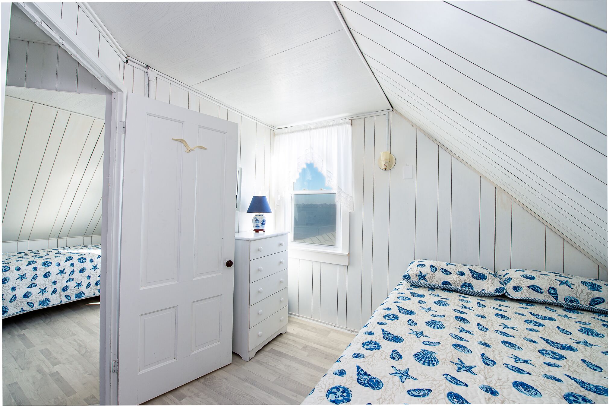A bedroom with a sloped ceiling, a white door, a dresser, a lamp, a window, and a bed with a patterned bedspread.