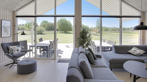Un vaste espace de vie avec une grande porte vitrée donnant sur une terrasse extérieure. À l’intérieur, on trouve un canapé gris, un pouf assorti et une table à manger avec des chaises.