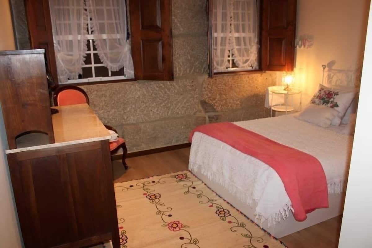 A bedroom with a bed, a desk, and a chair. There are two windows with curtains, a nightstand with a lamp, and a wall with a decorative pattern.