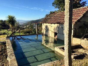 Uma piscina de pedra com água cristalina, cercada por paredes de pedra e uma estrutura de telhado em cerâmica.