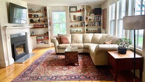 A living room with a beige sectional sofa, a fireplace, a large window, and a bookshelf filled with books and decorative items. - Waterfront, Swimming Hotspot, Private Down Town Location, Farmhouse (Waitsfield)