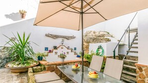 Una mesa de vidrio con sillas y una sombrilla, rodeada de macetas con plantas y un muro de piedra.
