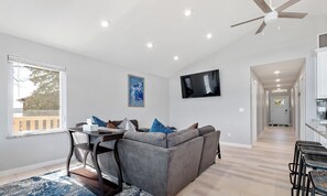 Een moderne woonkamer met een grijze modulaire bank, een eettafel met stoelen, een groot raam en een flatscreen televisie die aan de muur is bevestigd.