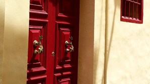Eine rote Tür mit Messingbeschlägen, in eine beige Wand eingesetzt, darüber ein kleines Fenster.
