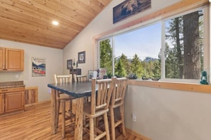 Un intérieur en bois avec une table et des chaises, une fenêtre donnant sur des arbres et des montagnes, et une pancarte de bienvenue.