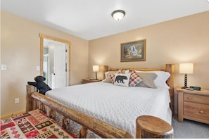 Une chambre à coucher avec un lit, des tables de chevet, une commode, un banc en bois, une chaise, une porte et un tableau au mur.