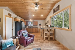 L’intérieur chaleureux d’un chalet comprend un coin repas, une cuisine et un fauteuil en cuir agrémenté d’un coussin orné d’un motif d’ours.