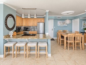 A kitchen with wooden cabinets, a stainless steel refrigerator, a granite countertop, and a dining area with wooden chairs. - Sabine 4D Waterfront Condo Sleeps 6, Corner Unit - Pool (Pensacola Beach)