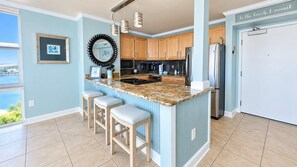 A kitchen with a granite island, wooden bar stools, a stainless steel refrigerator, and a view of the outdoors.