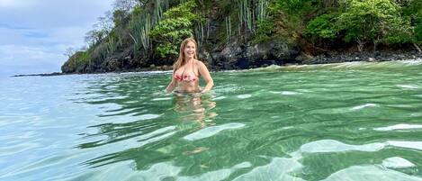 Ein klarer Blick auf einen tropischen Strand mit flachem Wasser, einer Person in einem Bikini und einem üppig bewachsenen, grünen Wald im Hintergrund.