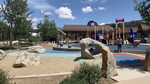 A playground with slides and climbing structures, surrounded by large rocks and trees. - Perfect Location! - EV charger, downtown, shuttle, + private hot tub & garage! (Winter Park)