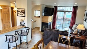 A modern living room with a fireplace, a dining area, and a kitchen with wooden cabinets and a bar. - Perfect Location! - EV charger, downtown, shuttle, + private hot tub & garage! (Winter Park)