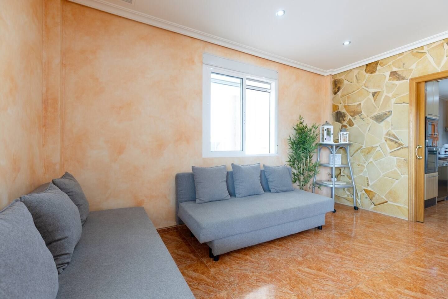 Un salon avec un canapé gris, un petit divan gris, une grande fenêtre, un mur en pierre et un coin cuisine visible à travers une porte ouverte.