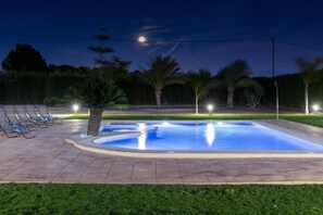 Piscina com deck de azulejos, cercada por palmeiras e uma fileira de cadeiras de descanso.