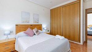 A hotel room with a wooden headboard, a bed with white bedding, a nightstand with a lamp, a wooden wardrobe, and a small bedside table with a plant. - Fidalsa Dom Perignon (Crevillente)